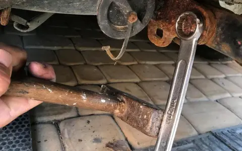 Zapieczona śruba poddawana próbie odkręcenia kluczem. Obok młotek i ręka.