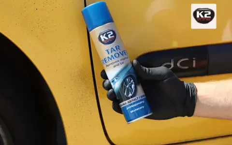 Rękawiczka z czarną gumą trzyma puszkę K2 Tar Remover, która skutecznie usuwa smołę z żółtego lakieru samochodu.