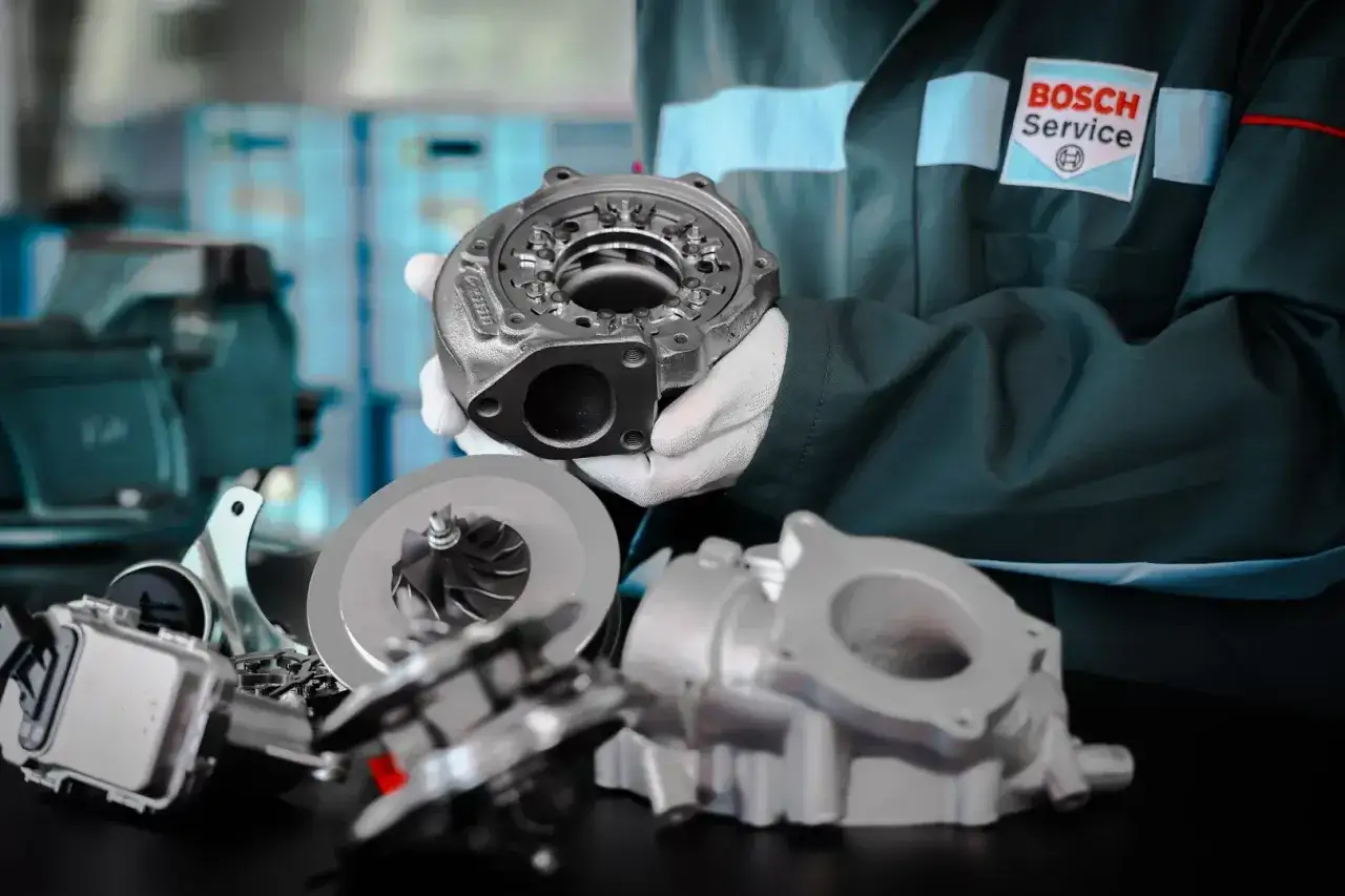 Mechanik w białych rękawiczkach prezentuje części turbosprężarki, ukazując jej skomplikowaną geometrię.