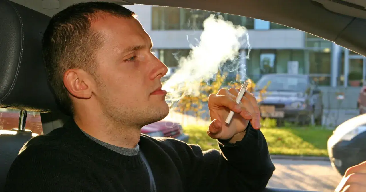 Mężczyzna pali papierosa w samochodzie, a dym unosi się w powietrzu. Zastanawiasz się, jak pozbyć się zapachu papieros&oacute;w z auta?